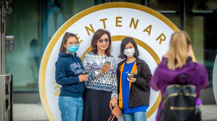 Three people holding cakes and posing for a picture in from of the centenary sign