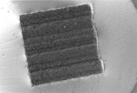 A micrograph of dark square that has a rough surface in a paler, smooth background