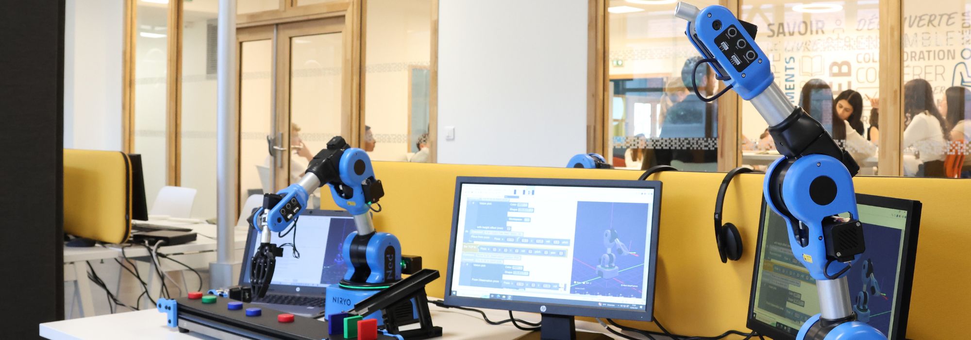 Two blue robotic arms are positioned on desks with computer monitors displaying coding software.