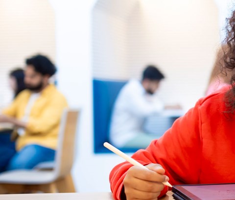 Student sitting at a table whilst consulting a book and writing on an IPad