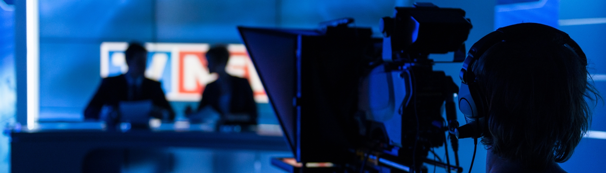 Production lit television studio (blue tones( with videographer in the foreground, facing camera to two broadcasters in a softly focussed background.