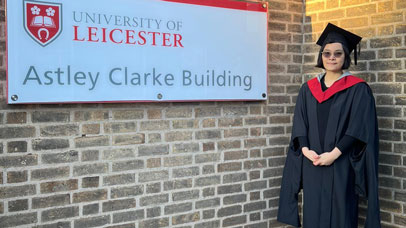 Student Frankie wearing graduation robes and standing next to a sign which reads: Astley Clarke Building