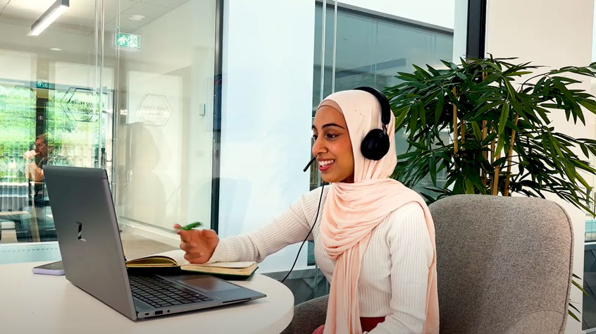 Student sitting at a table, speaking into a laptop and wearing a headset.