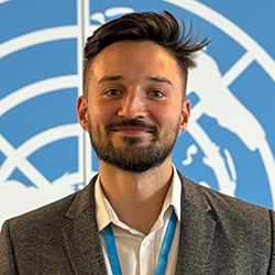 Aleksej Gubka, dressed in a white shirt and grey jacket, standing in front of a United Nations logo, smiling and looking directly at the camera