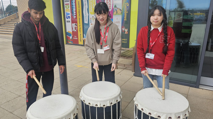Three people standing in front of traditional drums and holding sticks.