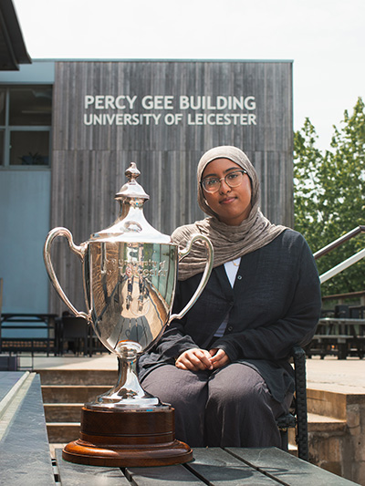 A woman sits on a bench, proudly holding a trophy, looking at the camera.