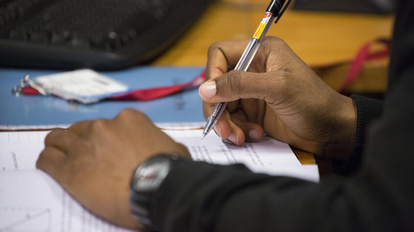 Close-up photograph of a student's hands, writing on a notebook with a biro