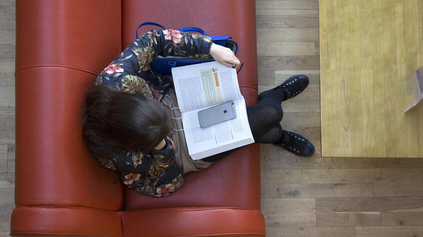 Overhead photo looking down on a woman reading a textbook, sat on a red sofa