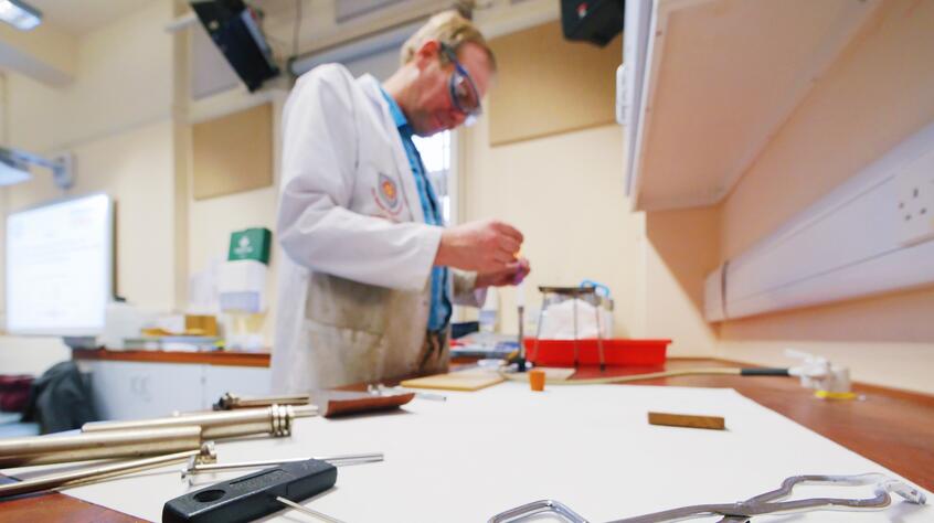 Photograph of a man in a lab coat and goggle working on something in a lab. Research notes and tongs are in the foreground.