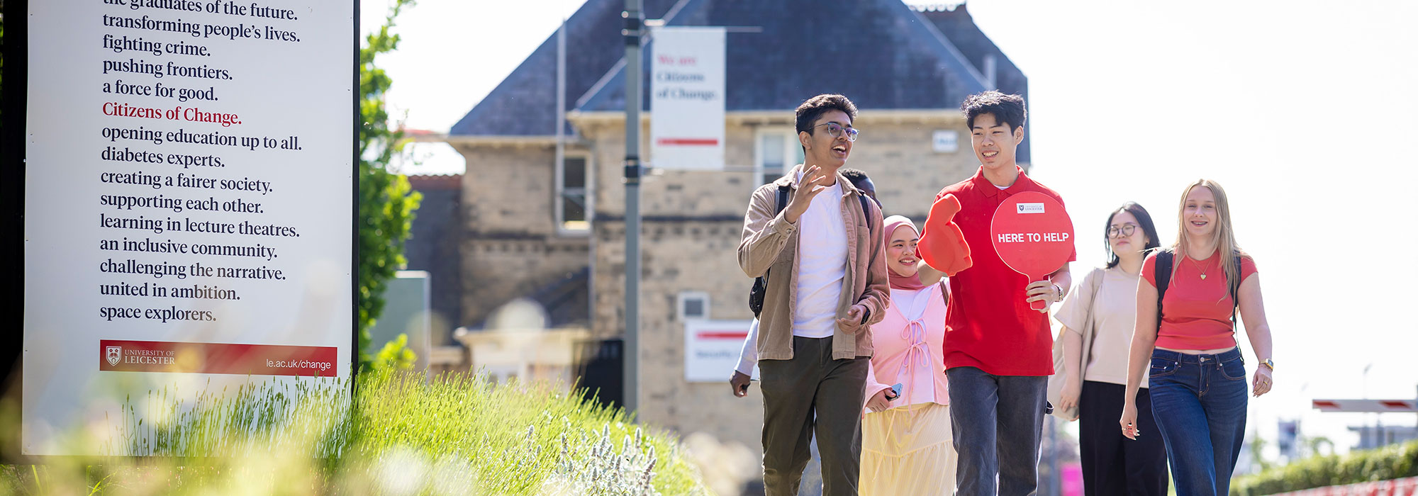 Group of prospective students walking along a landscaped campus path beside a large sign reading “Citizens of Change” with University of Leicester branding. One person in a red t‑shirt holds a speech‑bubble sign that says “HERE TO HELP,” while others walk alongside carrying bags. In the background are campus buildings, greenery, and additional signage.
