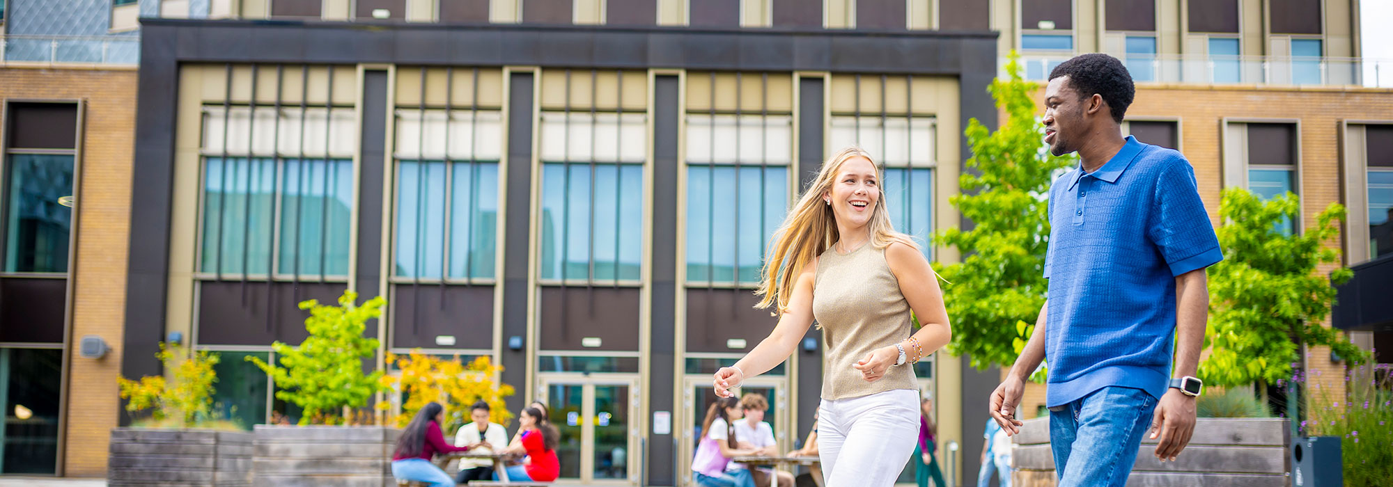 Two people walk side by side across an outdoor university-style courtyard, with a modern building featuring tall windows in the background. Wooden benches and tables are arranged in the space, where small groups of people sit and talk. One person wears a sleeveless light-colored top and light trousers, and the other wears a blue short-sleeved shirt and jeans. Trees, planters, and greenery frame the scene.