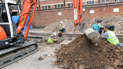 Archaeologists being filmed on an excavation site with a digger and large mound of earth in the foreground.