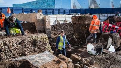 Number of archaeologists working at an excavation site with the Leicester Highcross in the background.