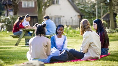 Three students sat on a bench in the background, with four students sat on a picnic blanket in the foreground, on a grassy area of campus.