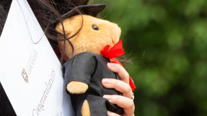Someone's hand holding both a teddy bear dressed in a black robe and mortarboard hat, and a certificate saying congratulations