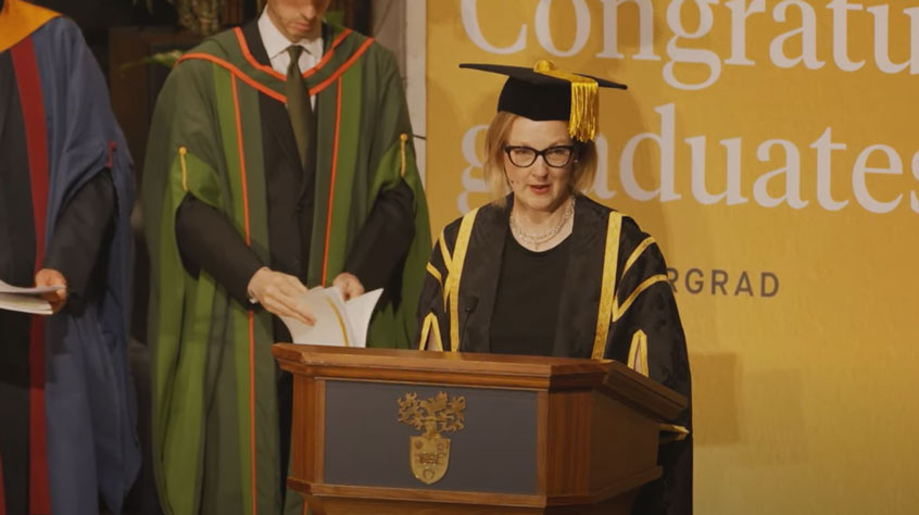 Henrietta O'Connor standing at a lectern in graduation robes.
