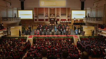 Wide view of a stage full of people and an audience of people in caps and gowns at graduation.