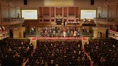 Wide view of a stage full of people and an audience of people in caps and gowns at graduation.