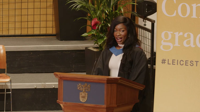 A woman dressed in a graduation gown giving a speech.