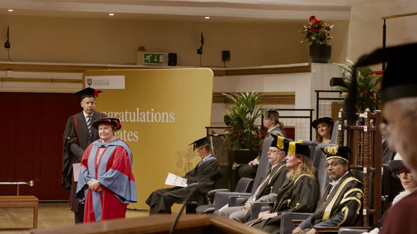 People dressed in graduation gowns, listening to the speaker. 