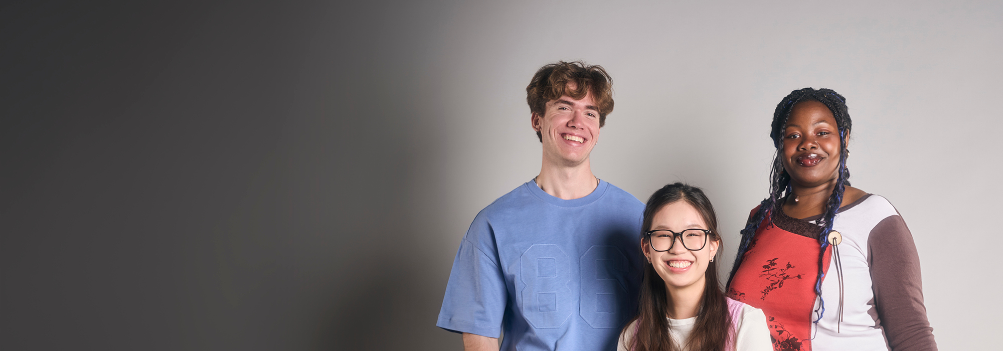 Three students smiling, with a grey background. 