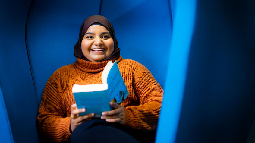 Student sitting in a big blue chair, reading a book and laughing.