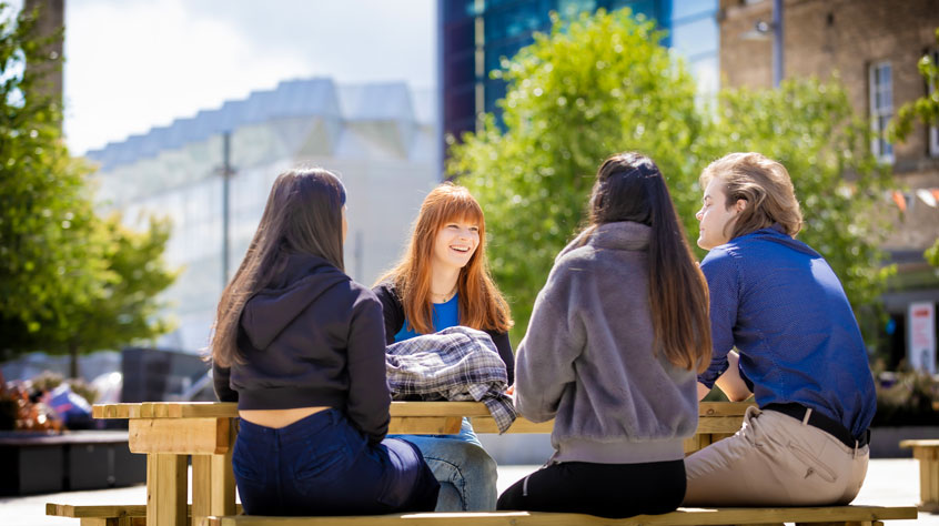 Students sitting on a bench talking, in Centenary Square on Leicester campus.