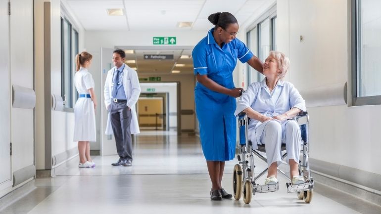 A nurse speaks to a smiling patient in a wheelchair in a hospital setting.