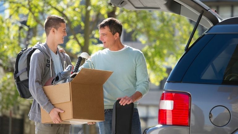 A student holding a cardboard box full of belongings is moved into University accommodation by a guardian.