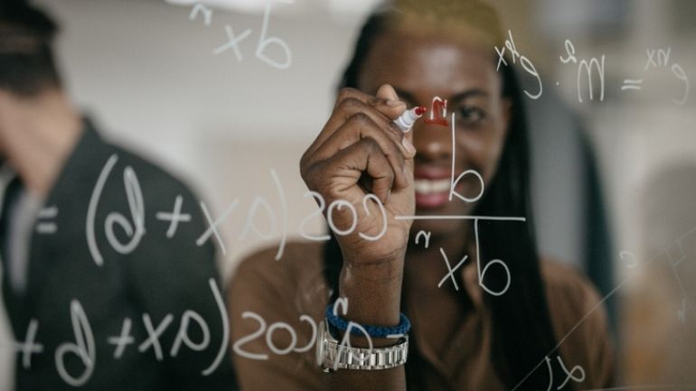 A Black female student writes mathematical equations on a transparent surface.