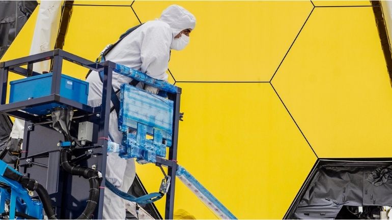 A technician wearing a protective suit and facemask inspects the gold-coated primary mirror of the James Webb Space Telescope.