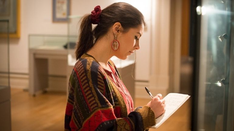 A young woman examines a museum exhibit and writes on a handheld notepad.