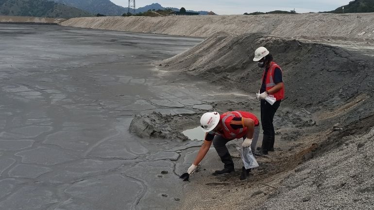 Two researchers in protective equipment sample a grey-brown pile of mine tailings.