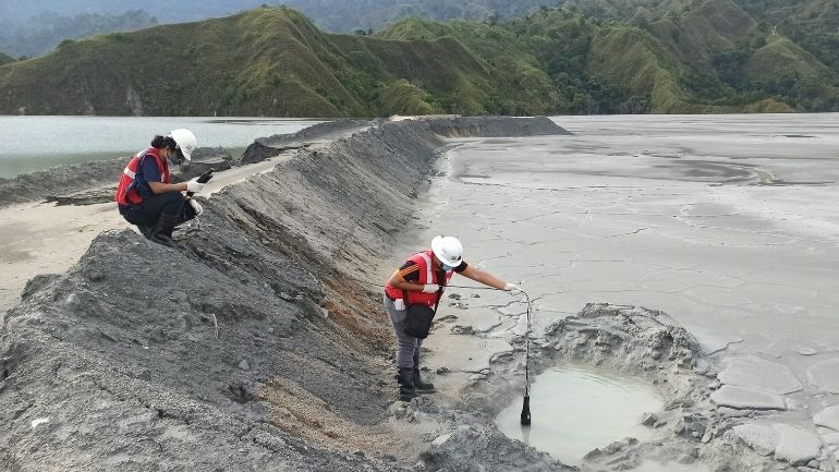 Two researchers in protective equipment sample a grey-brown pile of mine tailings.