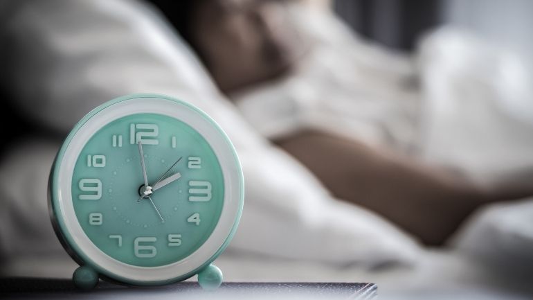 Close-up image of an alarm clock, with an individual in bed in the background.