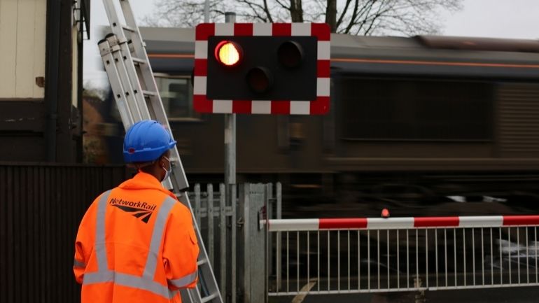 A Network Rail engineer stands in front of a red light at a level crossing as a train passes.