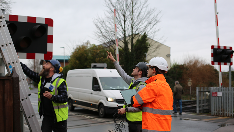 Workmen hold wiring and stand by a ladder near a level crossing wearing hardhats and high visibility jackets.