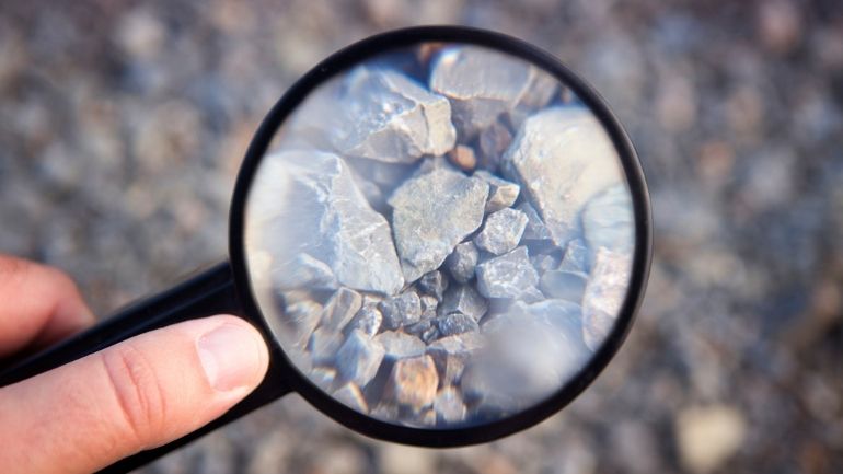 Close up of a hand holding a magnifying glass to study small rocks.