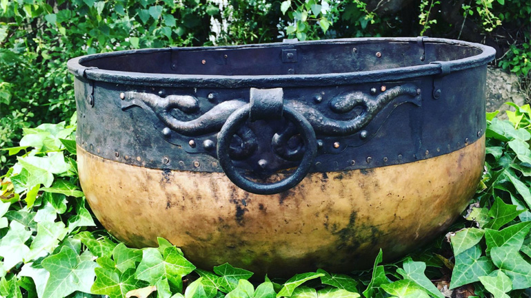 Reconstructed Glenfield Cauldron stitting on a bed of ivy leaves. The cauldren has a gold bottom and black top with an ornate handle attached.