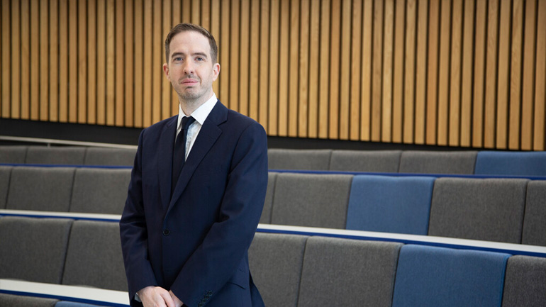 Professor Dan Ladley stands in an empty lecture theatre wiearing a navy suit looking into the camera