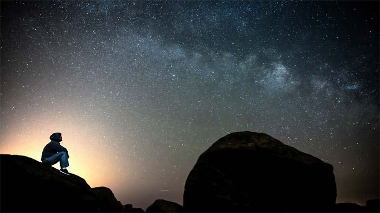 Man sitting on the rocks in solitude under the cover of dark, clear sky and looking at Milky Way galaxy while thinking about his existence