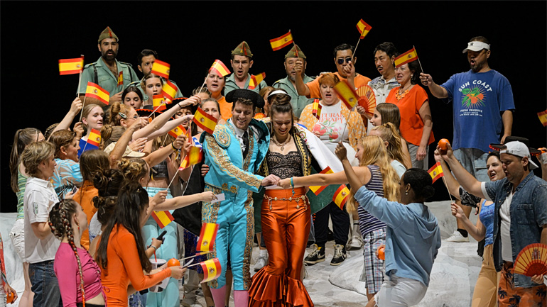 Liam James Karai dressed as a bull fighter whilst playing Escamillo in Carmen. She is surrounded by a crowd waving Spanish flags. 