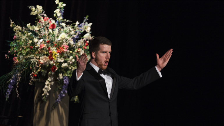 Liam James Karai wearing black suit, white shirt and bow tie singing with both hands raised in front of him. Large flower arrangement in background.