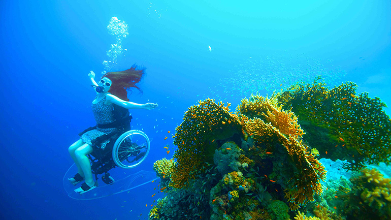 A woman in a wheelchair, wearing a swimming costume and scuba mask, underwater with arms stretched out. Coral is in the foreground. 