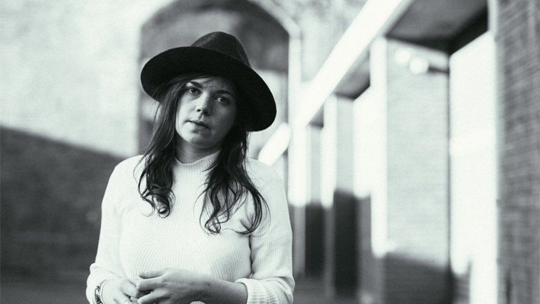 A black and white photo of Jessie Reid. She has long dark hair and is wearing a brimmed hat and white turtleneck. She is stood outside a brick industrial building.
