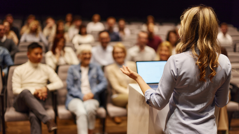 A woman with her back to the camera, presenting a lecture to full lecture theatre