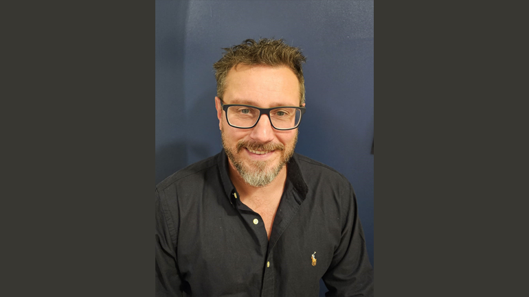 Andrew Futter wears a dark grey shirt and dark glasses whilst smiling at camera