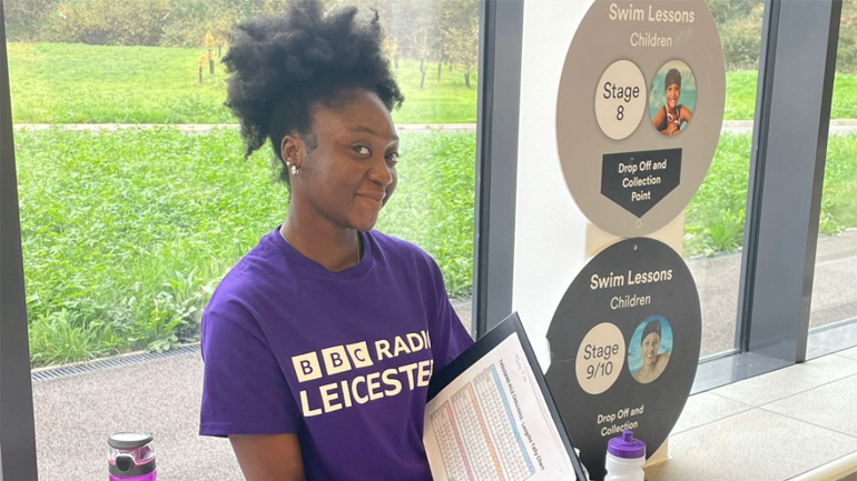 A University of Leicester student wears a purple BBC Radio Leicester tshirt and hold a clipboard at a swimming pool.