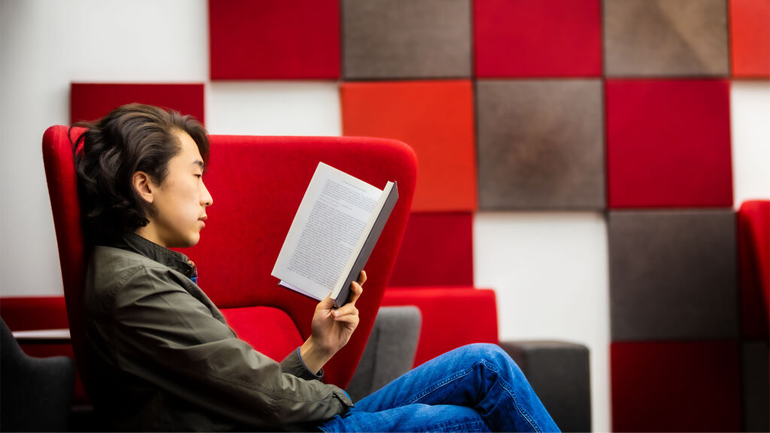 A male student reads a book in a red chair.