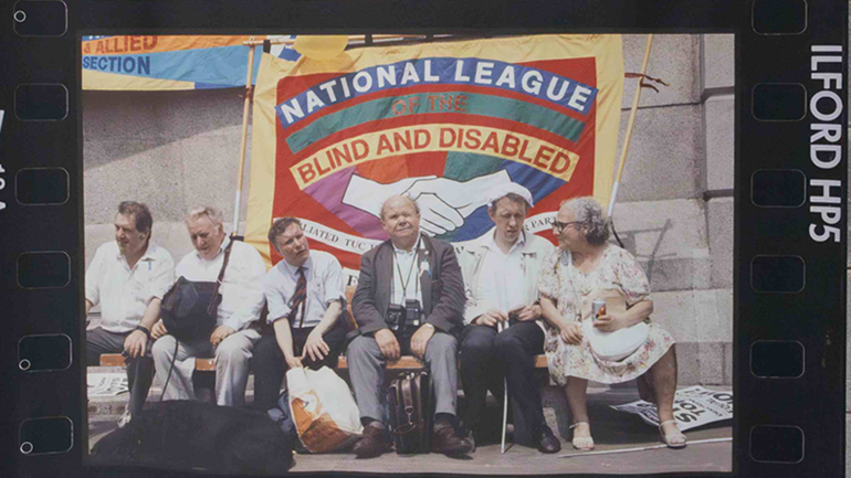 Six activists from the National League of the Blind and Disabled sit in front of a multicoloured fabric banner.